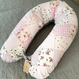 Könnte beinhalten: Ein rosa-weißes Patchwork-Schwangerschaftskissen mit Blumenmuster. Das Kissen ist U-förmig und hat eine graue Schleife.