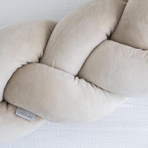 Op de afbeelding: Een gevlochten babybumper in een neutrale beige kleur. De bumper is gemaakt van zacht, pluche materiaal en is ontworpen om een veilige en comfortabele omgeving voor baby's te bieden. Het label leest "LOOLAY".