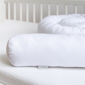 Op de afbeelding: Witte cilindrische kussen met een wit label met de tekst "LOOLAY" op een witte wiegmatras.