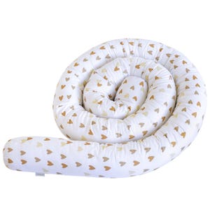Op de afbeelding: Een witte babybumper met een herhalend patroon van kleine, goudkleurige hartjes. De bumper is opgerold in een spiraalvorm, met een lang, recht gedeelte dat zich uitstrekt vanaf de spiraal. De bumper is ontworpen om een veilige en comfortabele omgeving voor baby's te bieden.