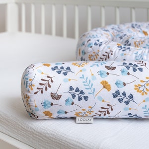 Op de afbeelding: Een wit, blauw en bruin baby nest met een witte achtergrond en een bloemenpatroon. Het nest is gemaakt van zachte stof en is ontworpen om een veilige en comfortabele slaapruimte voor een baby te bieden. Het nest is gelabeld met "LOOLAY".