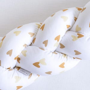 Op de afbeelding: Gevlochten wiegbumper van witte stof met een patroon van gouden hartjes. De bumper is gemaakt van zacht, gewatteerd materiaal en is ontworpen om de baby te beschermen tegen de harde oppervlakken van de wieg. Het product heeft een label met "LOOLAV".