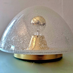 May include: A dome-shaped flush mount ceiling light featuring a clear glass shade with a textured bubble pattern. The fixture includes a central gold-toned metal socket housing a light bulb and a circular gold-toned metal base plate at the bottom.