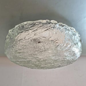 Könnte beinhalten: Eine kreisförmige Deckenleuchte mit einem dicken, klaren Glasschirm, der eine texturierte, gewellte Oberfläche aufweist, die das Aussehen von bewegtem Wasser nachahmt. Das Glas wird durch kleine Metallbeschläge an den Seiten der Leuchte befestigt.