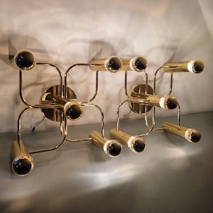 May include: Two brass wall sconces with a geometric design. Each sconce has multiple cylindrical light fixtures and spherical accents. The sconces are mounted on circular bases and have a modern, minimalist aesthetic.