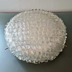 Könnte beinhalten: Eine runde, transparente Glasleuchte mit strukturierter Oberfläche und Gittermuster. Der Rand ist gezahnt. Zwei kleine Messingstäbe ragen aus den Seiten. Geeignet für Decken- oder Wandmontage.