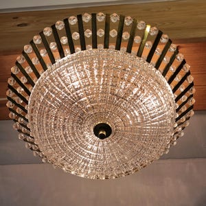 May include: A crystal chandelier with a circular design. The fixture features a large, textured glass globe at the center, surrounded by a ring of clear crystals and dark metal accents. The light fixture is mounted on a wooden ceiling.