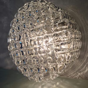 May include: A clear, spherical glass light fitting with a textured surface. The fitting is made up of many small, rounded glass elements, creating a bubbly, three-dimensional effect. Light reflects off the glass, creating highlights and shadows.