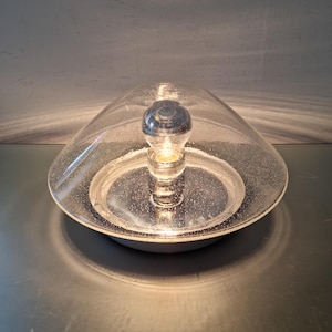 May include: A dome-shaped table lamp featuring a clear glass shade with a subtle speckled texture. The lamp has a central light bulb socket mounted on a circular, reflective silver-toned base that creates a bright, focused illumination effect.