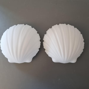 Könnte beinhalten: Zwei weiße, muschelförmige Dekoschalen. Jede Schale hat eine gerippte Textur, die von der Mitte ausstrahlt. Die Schalen werden auf einer grauen Oberfläche präsentiert.