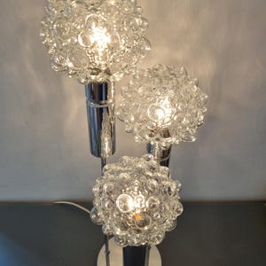 Puede incluir: Una lámpara de mesa de tres luces cromada con pantallas de vidrio de burbujas. La lámpara tiene un diseño moderno de mediados de siglo.
