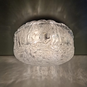 Könnte beinhalten: Eine Deckenleuchte aus mattiertem Glas. Die Leuchte hat ein strukturiertes, organisches Design und strahlt ein weiches, diffuses Licht aus. Die Leuchte ist rund und scheint aus dickem Glas zu bestehen.