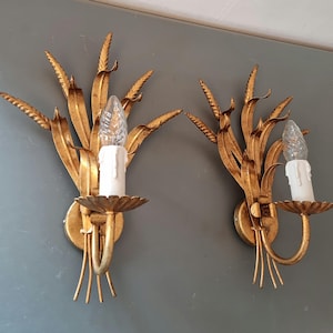 May include: A pair of gold-coloured wall sconces with a wheat stalk design. Each sconce has a white candle-shaped light bulb and a decorative candle holder. The sconces are mounted on a grey wall.