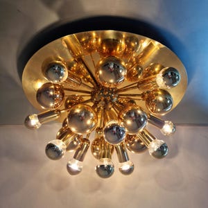 Könnte beinhalten: Eine goldfarbene Deckenleuchte im Sputnik-Stil. Die Leuchte hat eine kreisförmige Basis mit goldener Oberfläche, und mehrere kugelförmige Glühbirnen erstrecken sich nach außen. Die Glühbirnen sind eine Mischung aus Gold und Silber, was einen auffälligen Kontrast erzeugt. Die Leuchte ist an einer dunklen Decke montiert.