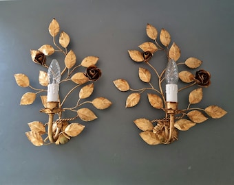 Peut inclure: Deux appliques murales dorées avec un motif floral. Chaque applique comprend un luminaire central avec une base blanche en forme de bougie et une ampoule transparente et pointue. Les appliques sont ornées de feuilles dorées et de roses.