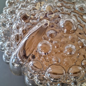 Könnte beinhalten: Nahaufnahme eines transparenten Glasobjekts mit einer blasenartigen, strukturierten Oberfläche. Das Glas hat einen warmen, bernsteinfarbenen Farbton im Hintergrund, wobei unterschiedlich große Blasen ein einzigartiges Muster bilden. Das Objekt scheint ein dekoratives Stück oder Teil eines größeren Designs zu sein.