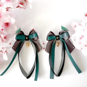 以下が含まれることがあります： 金色のアクセントと葉のチャームが付いた、緑と茶色のリボン2つのボウ。ボウは、花柄の白い生地に付けられています。