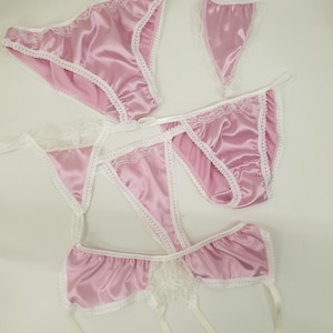 Pode incluir: Um conjunto de lingerie de cetim rosa com acabamentos em renda branca. O conjunto inclui uma cueca, um fio dental, uma cueca aberta e uma cinta-liga. A lingerie está disposta sobre uma superfície branca.