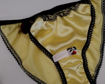 May include: Pale yellow satin panties featuring black lace trim along the edges, a thin black waistband, and a small dark red fabric rose accent on the left side. A small white rectangular tag is attached, displaying a rose image and text.