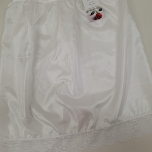 Può includere: Mezza sottoveste in raso bianco con un delicato bordo in pizzo sull'orlo. La sottoveste ha una vita arricciata e un'etichetta con testo e una grafica a rosa. Il tessuto ha una leggera lucentezza e il design generale è semplice ed elegante.