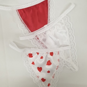 May include: Three satin thong underwear with white lace trim. One is red, one is white, and the third is white with red heart patterns. The underwear have thin straps and a triangular shape.