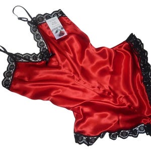 Può includere: Un completo composto da canotta e pantaloncini in raso rosso con bordo in pizzo nero lungo lo scollo e le aperture delle gambe. La canotta presenta sottili spalline regolabili e una piccola etichetta rettangolare bianca con una rosa.