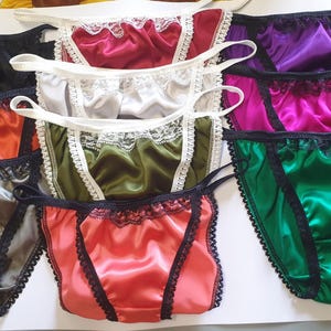 May include: A collection of satin underwear in various colors, including black, red, silver, olive green, coral, and purple. Each pair features delicate lace trim and thin straps for a comfortable fit. The garments are arranged on a white surface.