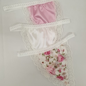 May include: Three pairs of lace trimmed underwear. One pair is pink satin, one is white satin, and one is white with a floral print.
