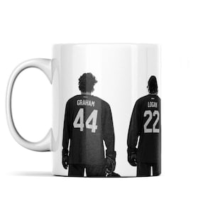 Puede incluir: Una taza de cerámica blanca con un gráfico en blanco y negro de dos jugadores de hockey vistos desde atrás. El jugador de la izquierda lleva una camiseta con el nombre GRAHAM y el número 44, mientras que el de la derecha lleva el nombre LOGAN y el 22.