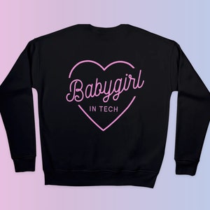 Op de afbeelding: Zwarte sweatshirt met een roze hartafbeelding en de tekst "Babygirl IN TECH" in een sierlijk lettertype. Het ontwerp is gecentreerd op de achterkant van de sweatshirt. De achtergrond heeft een roze en blauwe gradi&euml;nt.