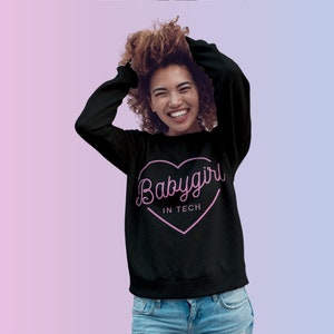 Op de afbeelding: Zwarte sweatshirt met een roze hartafbeelding met de tekst "Babygirl in Tech". De sweatshirt heeft lange mouwen en een ronde hals. De achtergrond is een verloop van roze en blauw.