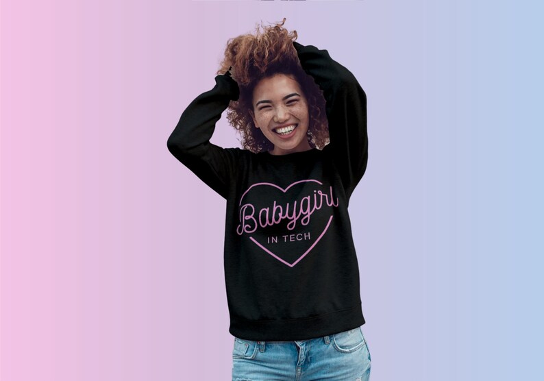 Op de afbeelding: Zwarte sweatshirt met een roze hartafbeelding met de tekst "Babygirl in Tech". De sweatshirt heeft lange mouwen en een ronde hals. De achtergrond is een verloop van roze en blauw.
