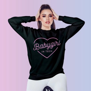Op de afbeelding: Zwart sweatshirt met lange mouwen met de tekst "Babygirl IN TECH" in een roze hart. Het sweatshirt wordt door een persoon gedragen. De achtergrond heeft een roze en blauwe gradi&euml;nt. Een casual kledingstuk.