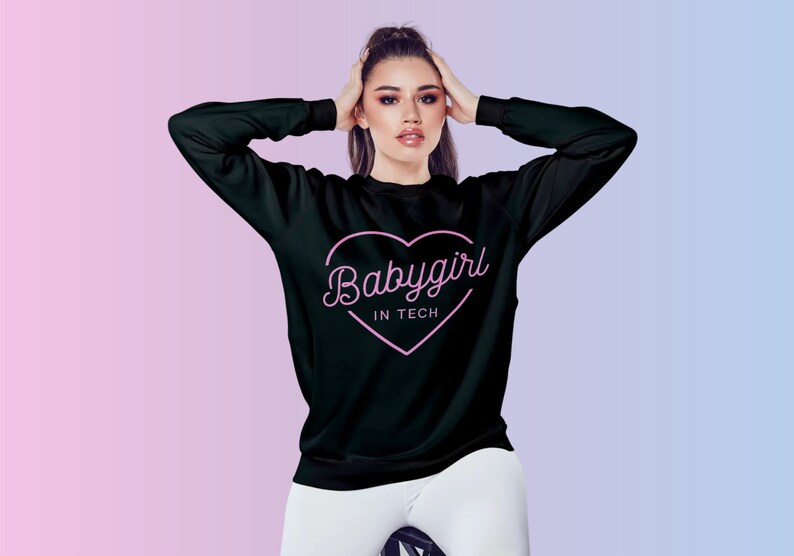 Op de afbeelding: Zwart sweatshirt met lange mouwen met de tekst "Babygirl IN TECH" in een roze hart. Het sweatshirt wordt door een persoon gedragen. De achtergrond heeft een roze en blauwe gradi&euml;nt. Een casual kledingstuk.