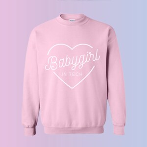 May include: A light pink sweatshirt with the words "Babygirl IN TECH" in a white heart-shaped design. The sweatshirt has a crew neck and long sleeves. The background has a gradient of pink and blue.