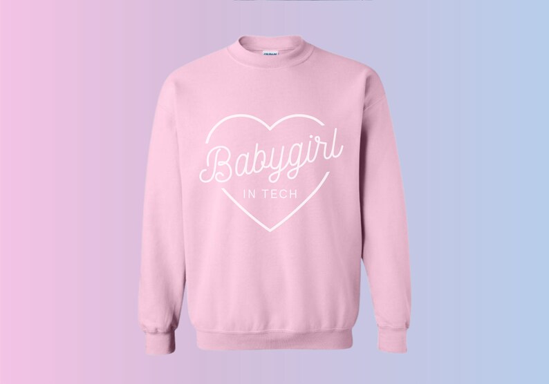May include: A light pink sweatshirt with the words "Babygirl IN TECH" in a white heart-shaped design. The sweatshirt has a crew neck and long sleeves. The background has a gradient of pink and blue.