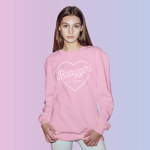 May include: A light pink sweatshirt with the text "Babygirl in Tech" inside a white heart. The long-sleeved top is worn by a person and is set against a pink and blue gradient background.