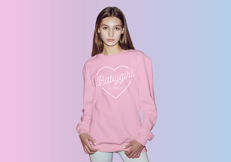May include: A light pink sweatshirt with the text "Babygirl in Tech" inside a white heart. The long-sleeved top is worn by a person and is set against a pink and blue gradient background.