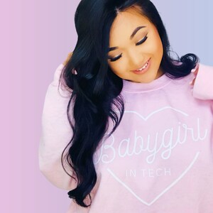 May include: A light pink sweatshirt with the text "Babygirl in Tech" inside a white heart. The sweatshirt is worn by a person with long, dark hair, against a pink and blue gradient background.