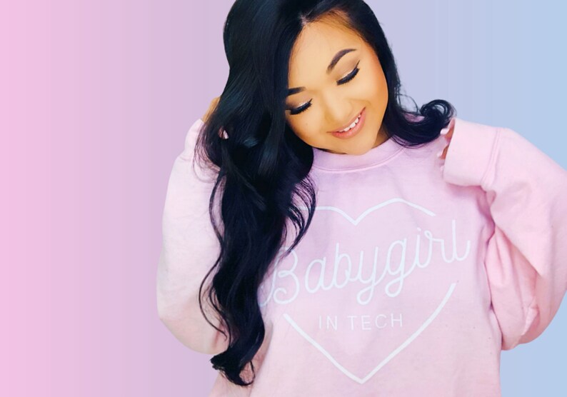 May include: A light pink sweatshirt with the text "Babygirl in Tech" inside a white heart. The sweatshirt is worn by a person with long, dark hair, against a pink and blue gradient background.