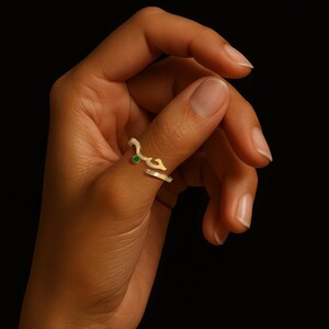 Puede incluir: Un anillo dorado con un diseño de escritura árabe y una pequeña gema verde. El anillo está en un dedo sobre un fondo negro. El diseño del anillo es elegante y moderno.