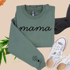 Könnte beinhalten: Ein grüner Pullover mit dem Wort "mama" in schwarzer, kursiv geschriebener Schrift auf der Vorderseite. Der Pullover hat ein kleines Herz und die Namen "Zack, Jeremy, Paityn, & Sam" auf dem linken Ärmel.