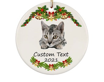 May include: A white ceramic Christmas ornament featuring a gray cat with yellow eyes. The ornament is decorated with a festive garland of holly, bells, and bows. Text on the ornament reads "Custom Text 2021".