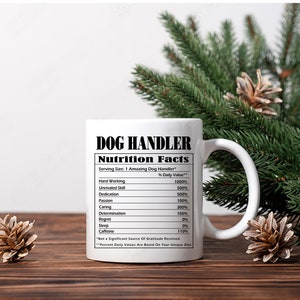 May include: White ceramic mug with the text "DOG HANDLER" and "Nutrition Facts". The nutrition facts list qualities like "Hard Working" and "Unrivaled Skill". The mug is on a wooden surface with pine cones and greenery.