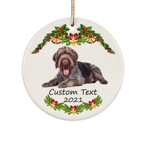 May include: A round ceramic Christmas ornament featuring a brown and white German Wirehaired Pointer dog lying down. The dog is surrounded by a green garland with red bows, gold bells, and red berries. The text "Custom Text 2021" is printed below the dog.