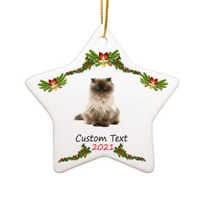 May include: A white ceramic star-shaped Christmas ornament with a brown and white cat sitting in the center. The ornament is decorated with green holly and red berries, and gold bells. The text "Custom Text 2021" is printed on the ornament.