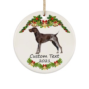May include: A white ceramic Christmas ornament featuring a brown and white dog, surrounded by festive greenery, bells, and berries. The ornament has the text "Custom Text 2021" in a decorative font. A gold string is attached for hanging.