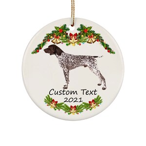 May include: White ceramic Christmas ornament featuring a German Shorthaired Pointer dog with brown and white markings. The ornament is decorated with festive greenery, bells, and the text "Custom Text 2021". A gold string is attached for hanging.