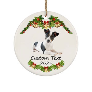 May include: White and black Jack Russell Terrier dog laying down on a white ceramic Christmas ornament. The ornament is decorated with a green garland with red berries, gold bells, and red bows. The text "Custom Text 2021" is printed on the ornament.