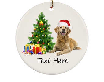 Puede incluir: Adorno navideño redondo con un Golden Retriever con gorro de Papá Noel, un árbol de Navidad decorado y regalos envueltos. El adorno tiene una cuerda dorada para colgar y el texto "Text Here" en la parte inferior.